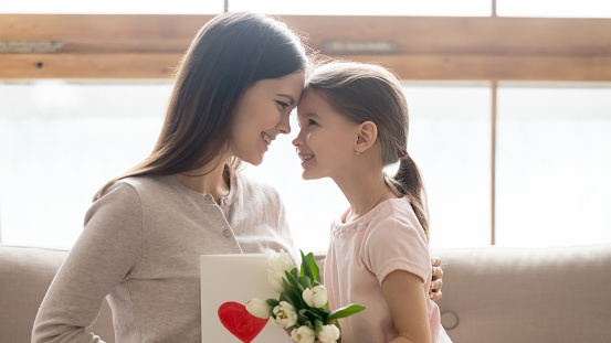 Happy mother and little daughter touching foreheads, thanking for gift, cute preschool girl congratulating mum with birthday or mothers day, presenting flowers and postcard, horizontal banner