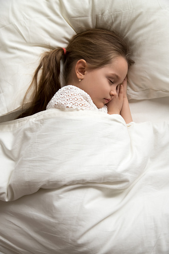 Calm adorable child lying on soft pillows, taking day nap under warm blanket, preschool girl sleeping alone in fresh comfortable bed with white linens, healthy peaceful sleep, vertical photo