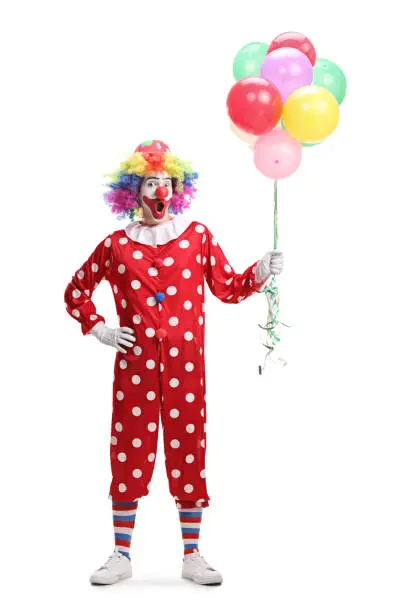 Full length portrait of a cheerful clown holding a bunch of balloons isolated on white background