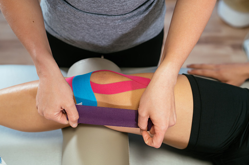 Photo detail of the hands of a physiotherapist woman gluing purple medical tape on another celestial tape and another pink one on the knee of a patient. Concept of muscle health and relaxation. Close-up