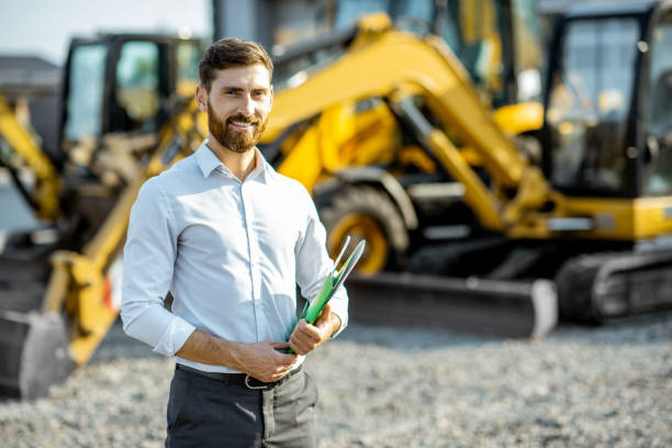 Salesman at the shop with heavy machinery Portrait of a handsome sales consultant or manager standing on the open ground of the shop with heavy machinery for construction agricultural machinery stock pictures, royalty-free photos & images