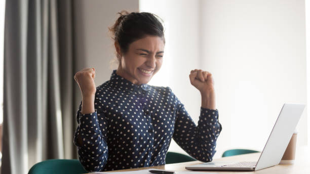 Excited happy indian employee celebrating success at work Happy indian woman office worker feeling excitement raising fists celebrates career ladder promotion or reward, businesswoman sitting at desk receive online news, great results successful work concept sales win stock pictures, royalty-free photos & images
