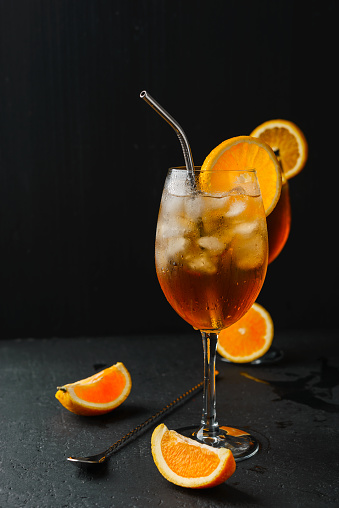 Alcoholic cocktail Spritz in a wine glass covered with water drops, slice of orange ,ice cubes and metal straw on dark background