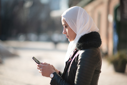 A beautiful young Muslim woman wearing a hijab and dressed in winter clothing is standing outside. She is texting on a mobile phone. The sun is shining on her face. The woman has a contemplative expression and is standing profile to the camera.