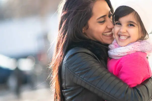 A beautiful young Muslim mother bonds with her daughter one winter afternoon. They are dressed warmly and are smiling while the mother embraces the girl in a loving hug.