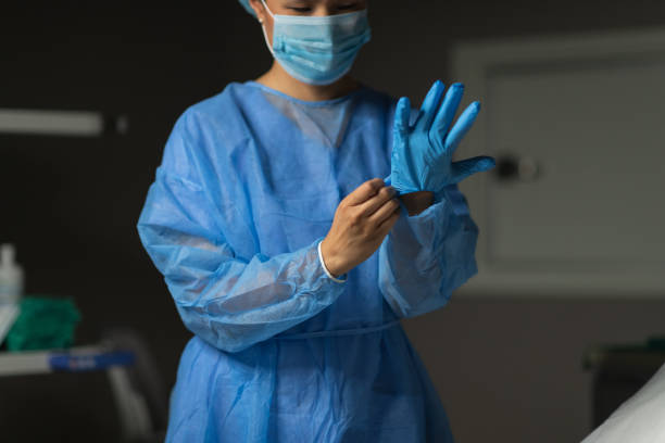 Female surgeon prepares for medical operation A female surgeon of Chinese descent is preparing for operation. She is standing in a hospital operating room next to an operating table. The doctor is dressed in a surgical gown and cap. She is putting on surgical gloves. putting on latex gloves stock pictures, royalty-free photos & images