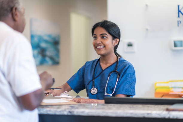 Medical professional greets patient at clinic A young female nurse of Indian descent greets a male senior patient. The medical professional is at the reception desk at a medical clinic. She is smiling kindly at the black man while checking him in for his appointment. receptionist stock pictures, royalty-free photos & images