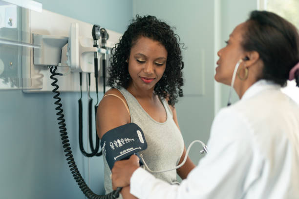 Young woman in medical consultation with female doctor A young black woman is at a routine medical appointment. Her healthcare provider is an ethnic woman. The patient is sitting on an examination table in a clinic. The doctor is checking the patient's blood pressure. The patient is smiling while looking down at her arm. blood pressure medication stock pictures, royalty-free photos & images