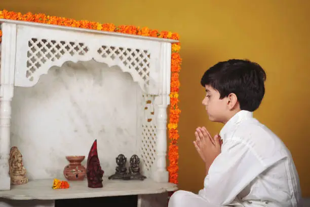 Photo of One praying Indian Asian boy doing Pooja or prayer in front of a marble temple mandir with idols of Lord Ganesh and Goddess Lakshmi, with marigold flowers