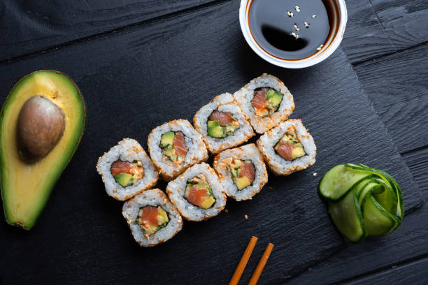 set di involtini di sushi con crema di formaggio, riso e salmone su una tavola nera decorata con salsa di soia e avocado su uno sfondo di legno scuro. cucina giapponese. sfondo della foto del cibo - prepared tuna foto e immagini stock