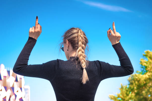 eine junge schlanke frau zeigt den mittelfinger mit zwei händen, dreht den rücken, der blaue himmel im hintergrund. das mädchen schickt alle, um sie zuficken symbol senden sie! - fuck you stock-fotos und bilder