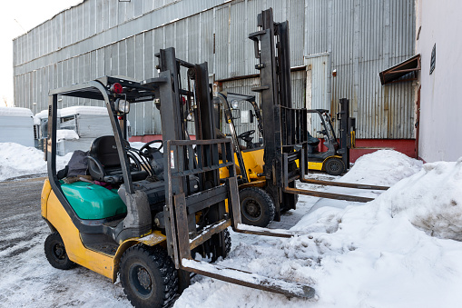 Several forklifts stand near the warehouse building. Winter snow day.
