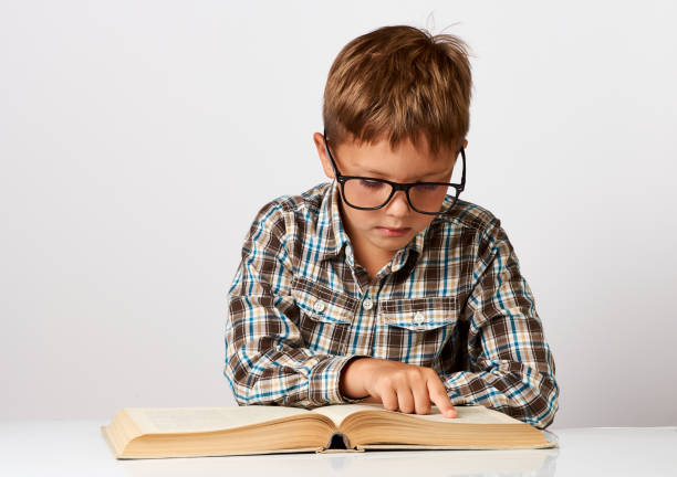 el niño lindo está sentado en el escritorio con el libro. niño de siete años - eyewear child glasses 6 7 years fotografías e imágenes de stock