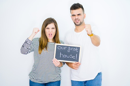 Young couple holding blackboard with our first home text over white isolated background annoyed and frustrated shouting with anger, crazy and yelling with raised hand, anger concept