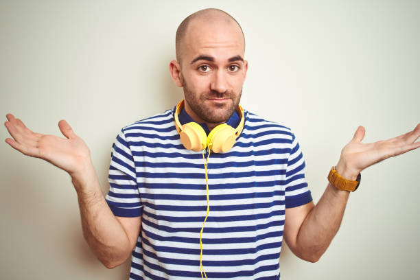 young man listening to music wearing yellow headphones over isolated background clueless and confused expression with arms and hands raised. doubt concept. - head and shoulders audio imagens e fotografias de stock