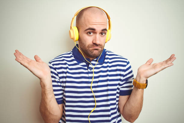 young man listening to music wearing yellow headphones over isolated background clueless and confused expression with arms and hands raised. doubt concept. - head and shoulders audio imagens e fotografias de stock