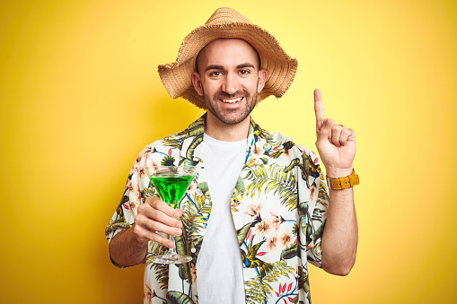 Young man wearing summer hawaiian flowers shirt and drinking a cocktail over yellow background surprised with an idea or question pointing finger with happy face, number one