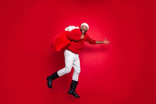 Full body profile side photo of cheerful guy in xmas santa claus hat holding carrying, bag in christmas night wearing pullover pants trousers boot isolated over red background