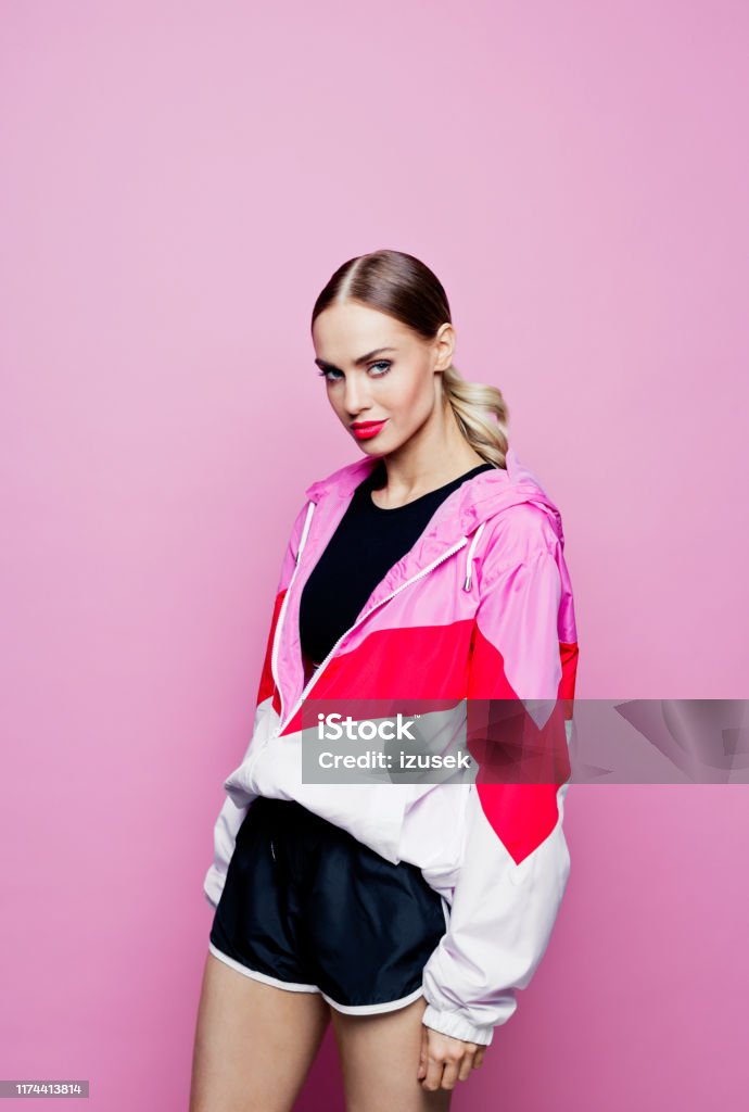 80's style portrait of beautiful woman in oversized tracksuit against pink background Mid adult beautiful woman wearing sport clothes and shorts standing against pink background, looking at camera. Fashion Stock Photo