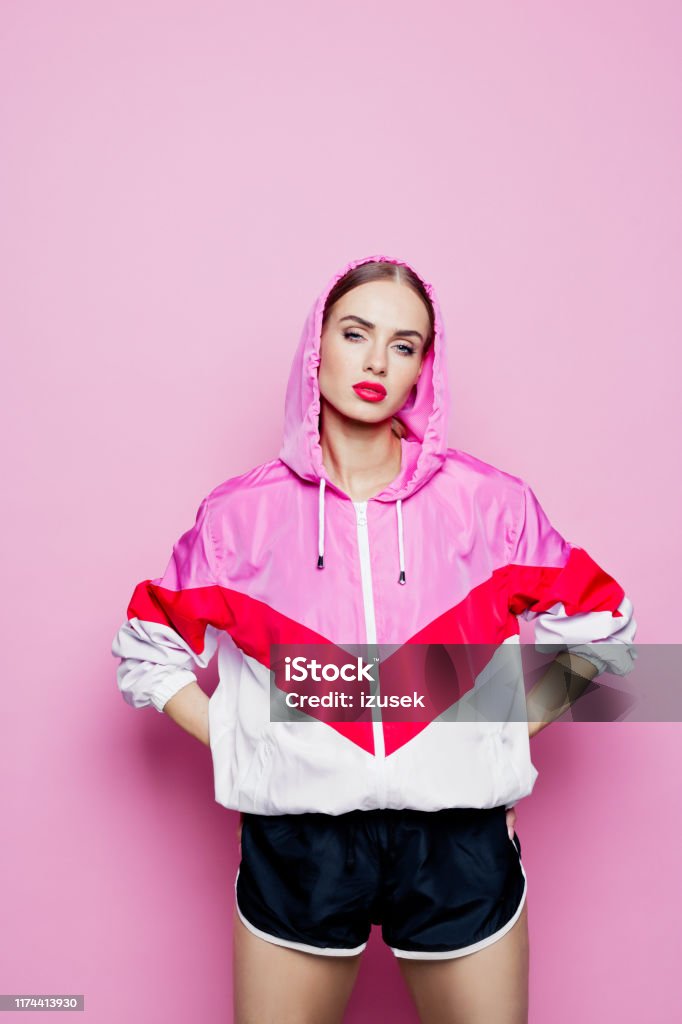 80's style portrait of beautiful woman in oversized tracksuit against pink background Mid adult beautiful woman wearing oversized tracksuit and shorts standing against pink background, looking at camera. Women Stock Photo