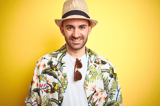 Young man on vacation wearing hawaiian flowers shirt and summer hat over yellow background with a happy and cool smile on face. Lucky person.