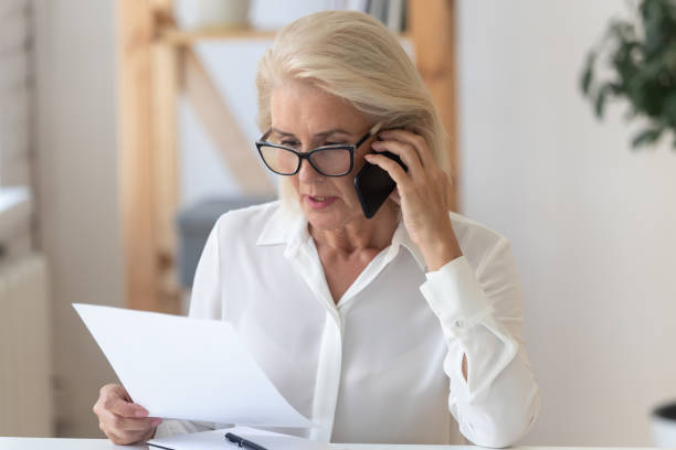 Serious senior woman talk on cellphone discussing paperwork