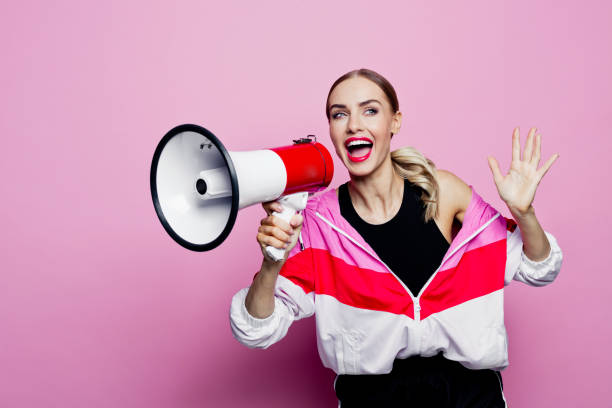 80's style portrait of happy woman in sports clothes holding megaphone Mid adult beautiful woman wearing oversized tracksuit and shorts standing against pink background, holding megaphone in hands and smiling at camera. announce stock pictures, royalty-free photos & images