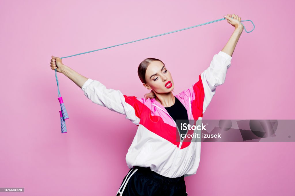 80's style portrait of smiling in sports clothes against pink background Mid adult beautiful woman wearing oversized tracksuit and shorts standing against pink background, holding jumping rope above her head. Sport Stock Photo