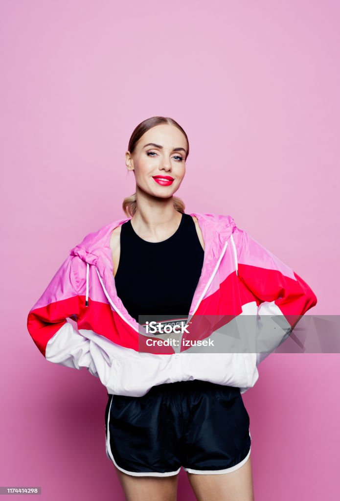 80's style portrait of smiling woman in sports clothes against pink background Mid adult beautiful woman wearing oversized tracksuit and shorts standing against pink background, looking at camera. 30-34 Years Stock Photo