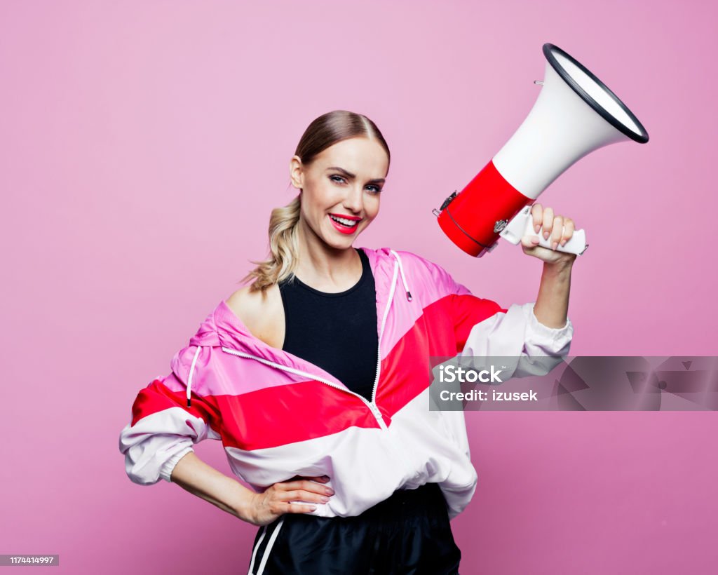 80's style portrait of smiling woman in sports clothes holding magaphone Mid adult beautiful woman wearing oversized tracksuit and shorts standing against pink background, holding megaphone in hands and smiling at camera. Megaphone Stock Photo