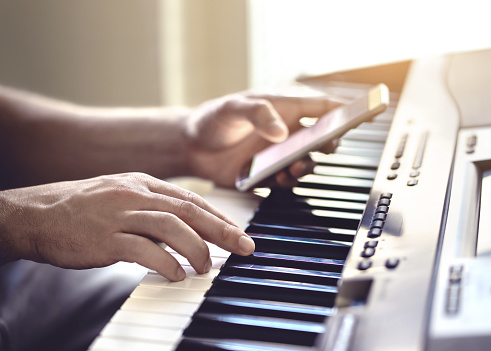 Man playing piano and using mobile phone. Person recording sound, reading notes from smartphone screen or writing lyrics for a song. Pianist watching online tutorial video and lesson and practising.