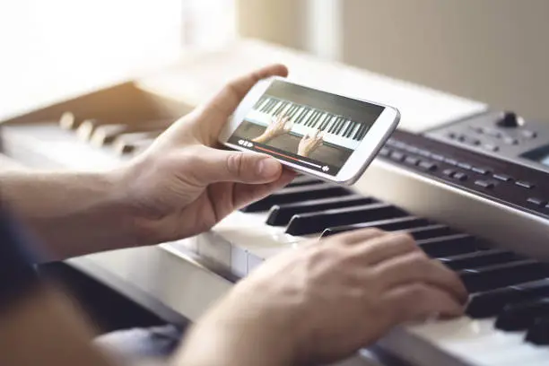 Photo of Piano lesson online. Man watching video tutorial with mobile phone and practising playing. Person learning to play an instrument with an internet course and class.
