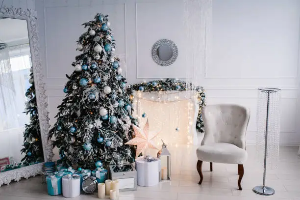 Decorated Christmas interior. Christmas tree with gifts boxes in a white room. Fir-tree, armchair, fireplace decorated with garlands. Decor. Happy New Year and Merry Christmas.