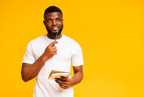 Pensive african american millennial man taking notes, writing checklist in notebook, looking upwards deep in his minds. Yellow background with free space