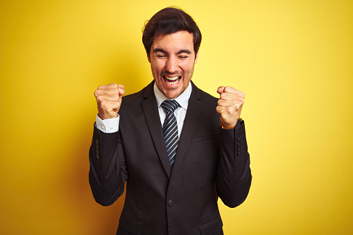 Young handsome businessman wearing suit and tie standing over isolated yellow background excited for success with arms raised and eyes closed celebrating victory smiling. Winner concept.