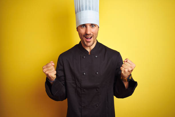 istock Young chef man wearing uniform and hat standing over isolated yellow background celebrating surprised and amazed for success with arms raised and open eyes. Winner concept. 1174438986