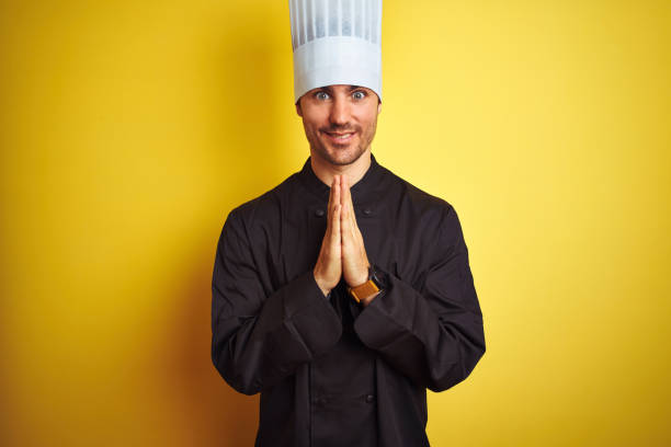 istock Young chef man wearing uniform and hat standing over isolated yellow background praying with hands together asking for forgiveness smiling confident. 1174439023