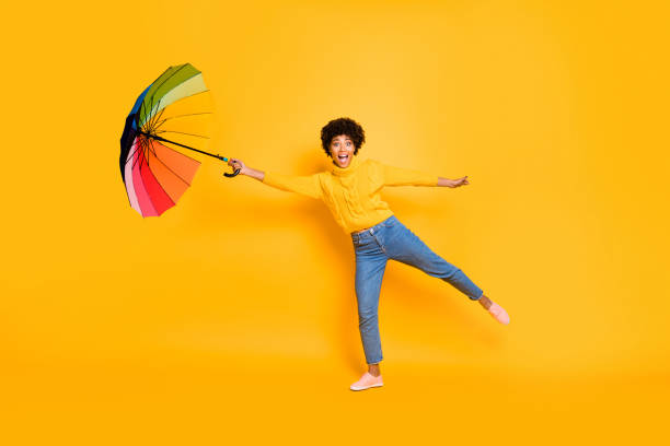 ホールド・オン・アドベンチャーズは、傘を手に持ち、風の孤立した背景に吹き飛ばされようとしないジーンズの人々に驚いた興奮した陽気なショックを受けたファンキーのフルボディ写真� - umbrella women storm yellow ストックフォトと画像