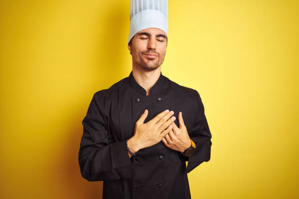 istock Young chef man wearing uniform and hat standing over isolated yellow background smiling with hands on chest with closed eyes and grateful gesture on face. Health concept. 1174439512