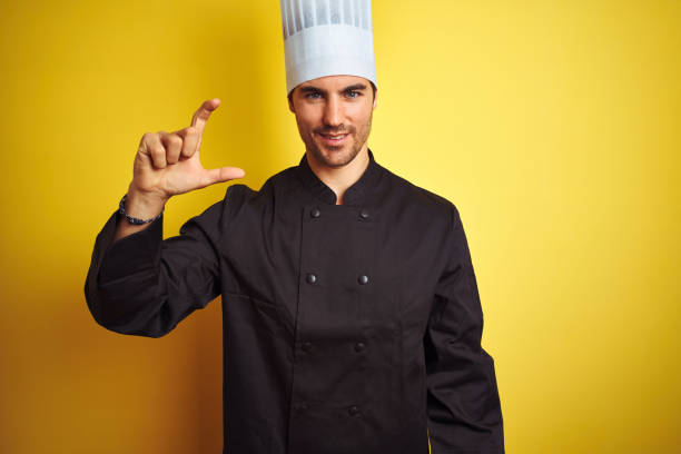 istock Young chef man wearing uniform and hat standing over isolated yellow background smiling and confident gesturing with hand doing small size sign with fingers looking and the camera. Measure concept. 1174439579