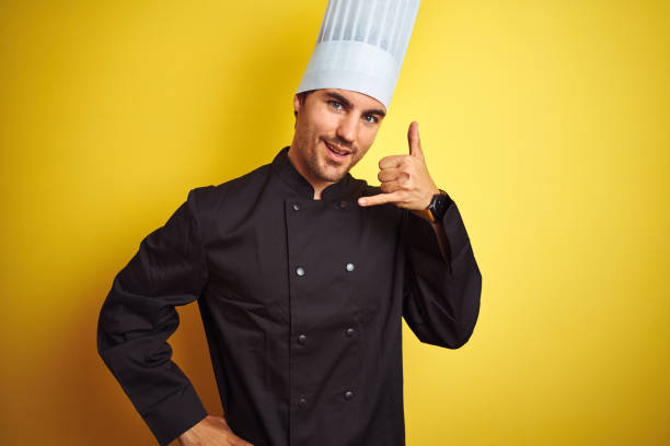 istock Young chef man wearing uniform and hat standing over isolated yellow background smiling doing phone gesture with hand and fingers like talking on the telephone. Communicating concepts. 1174439606