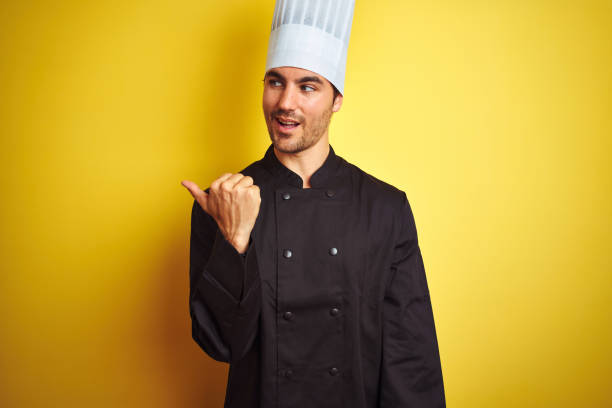 istock Young chef man wearing uniform and hat standing over isolated yellow background smiling with happy face looking and pointing to the side with thumb up. 1174439735