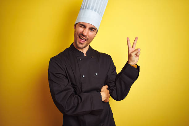 istock Young chef man wearing uniform and hat standing over isolated yellow background smiling with happy face winking at the camera doing victory sign. Number two. 1174439777