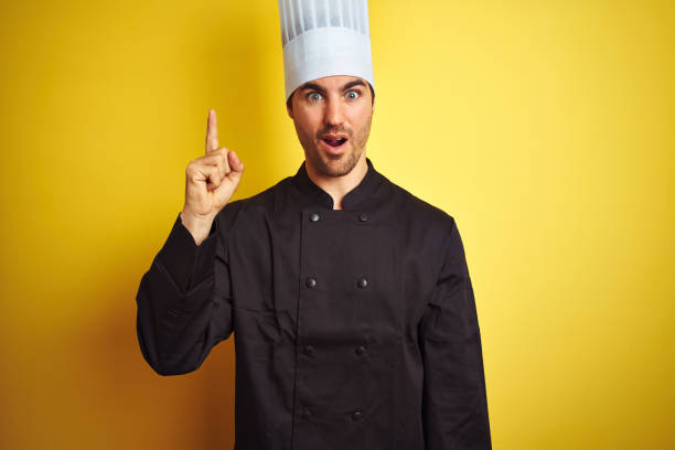 istock Young chef man wearing uniform and hat standing over isolated yellow background pointing finger up with successful idea. Exited and happy. Number one. 1174439865