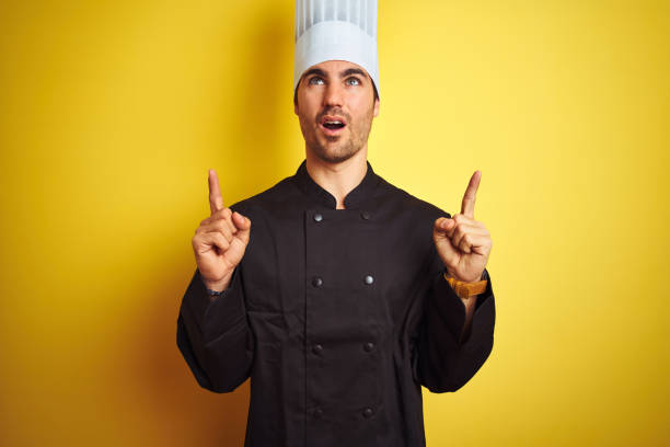 istock Young chef man wearing uniform and hat standing over isolated yellow background amazed and surprised looking up and pointing with fingers and raised arms. 1174439872