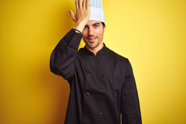 Young chef man wearing uniform and hat standing over isolated yellow background surprised with hand on head for mistake, remember error. Forgot, bad memory concept.