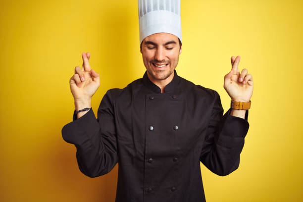 istock Young chef man wearing uniform and hat standing over isolated yellow background gesturing finger crossed smiling with hope and eyes closed. Luck and superstitious concept. 1174439906