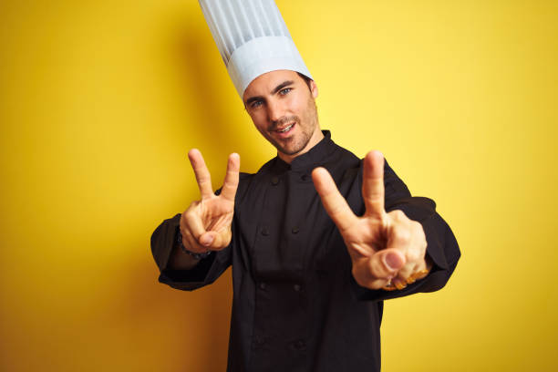 istock Young chef man wearing uniform and hat standing over isolated yellow background smiling looking to the camera showing fingers doing victory sign. Number two. 1174439942
