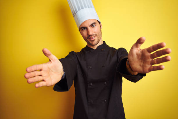istock Young chef man wearing uniform and hat standing over isolated yellow background looking at the camera smiling with open arms for hug. Cheerful expression embracing happiness. 1174439967