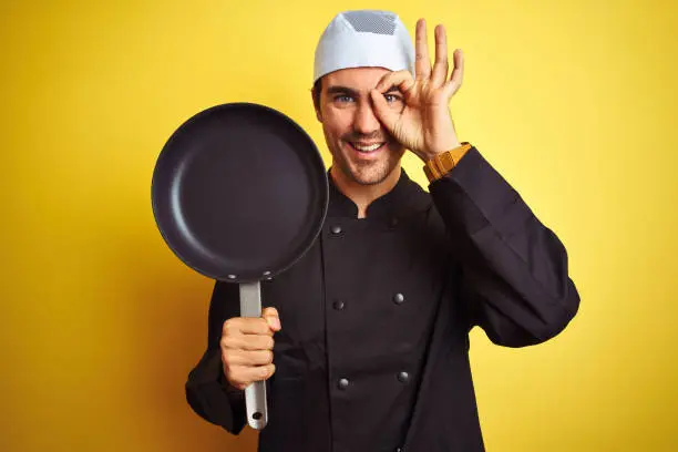 Photo of Young chef man wearing uniform and hat holding cook pan over isolated yellow background with happy face smiling doing ok sign with hand on eye looking through fingers